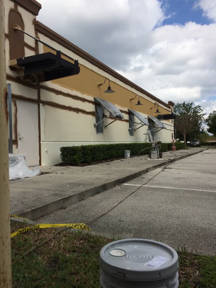 A long storefront wall half repainted in cream and gold, awnings wrapped in plastic, sprayer and buckets below.