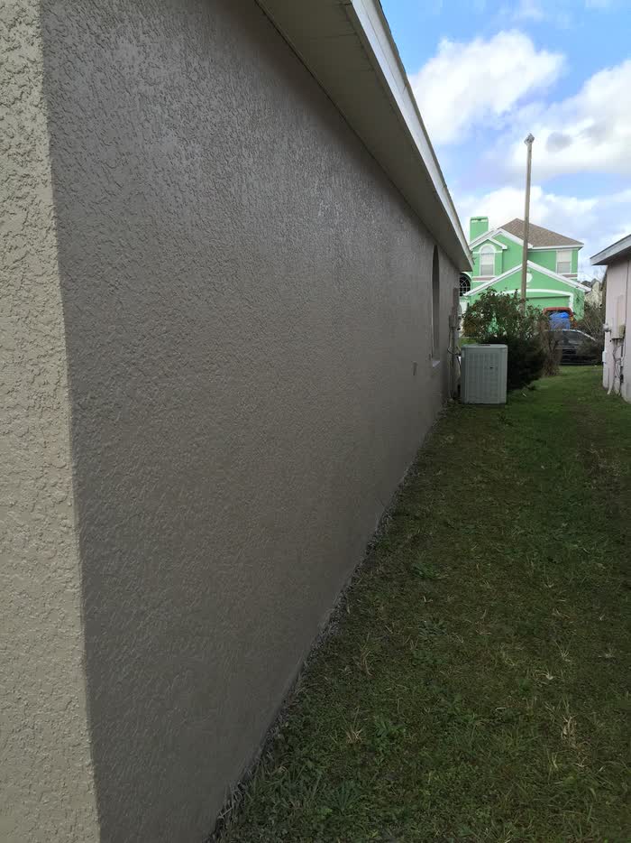 Grey side wall of a house running back to an air conditioning unit, green house next door.