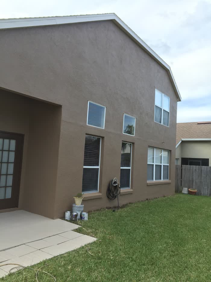 Side wall of a two-storey taupe house, with paint tins and a coiled hose on the grass.
