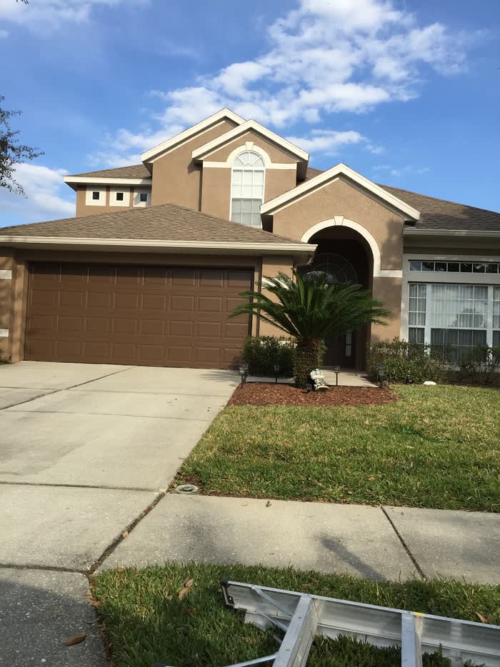 A two-story house in tan with cream trim and a brown double garage door, a ladder lying on the grass.