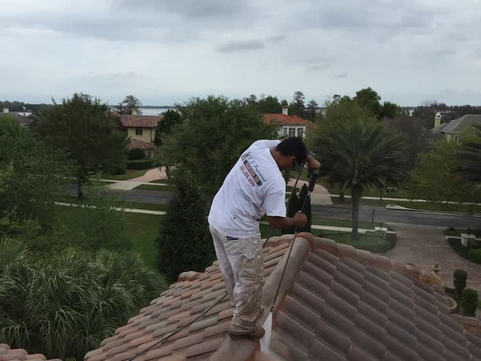 A worker steadies a long spray wand along the ridge of a curved-tile roof above a leafy street.