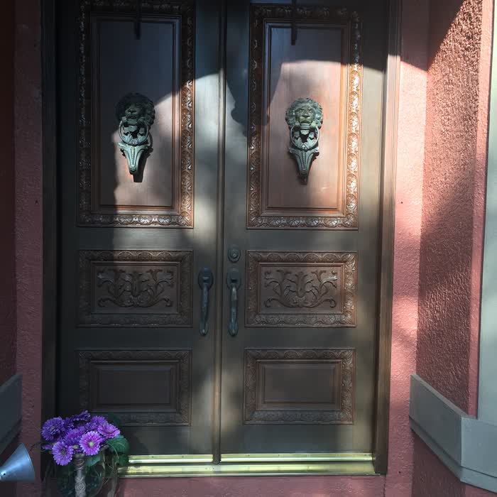 Ornate dark double doors with lion-head knockers set in a pink wall, purple flowers beside the step.