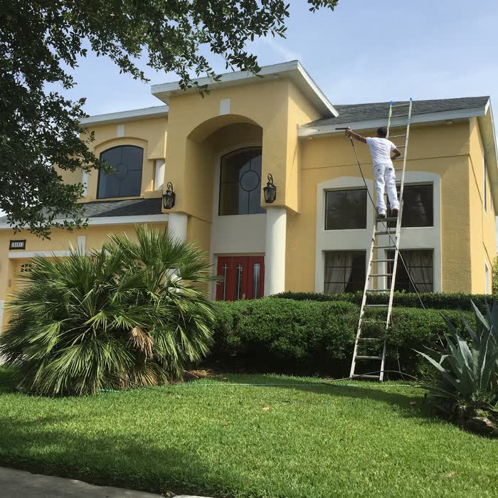 A painter high on a ladder works along the white trim of a yellow house with red front doors.