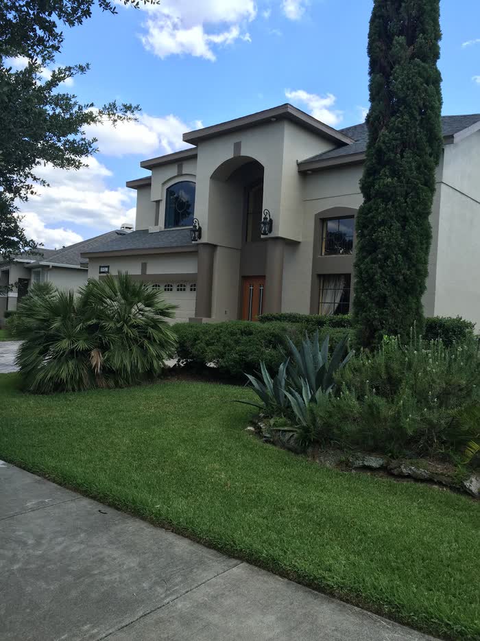 Taupe two-story house seen past a tall cypress, with palms and spiky agave filling the front bed.