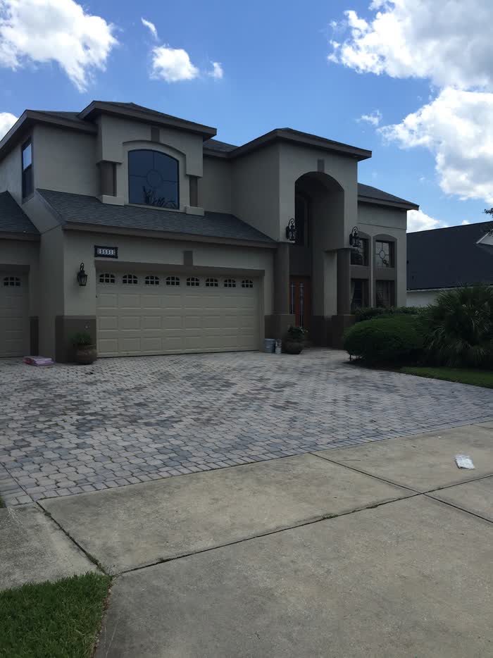 Wide grey-beige house with cream garage doors, a paver driveway out front and buckets set by the wall.