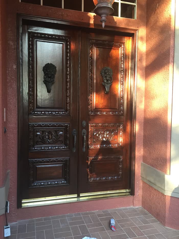 Deep reddish-brown double doors with carved panels and lion-head knockers, warm in low sun against a coral wall.