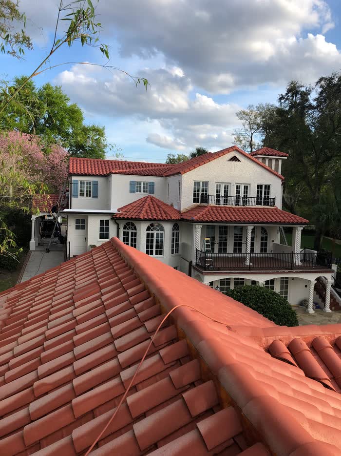 Seen from a rooftop, red barrel tiles run toward a white two-storey house with iron balconies.