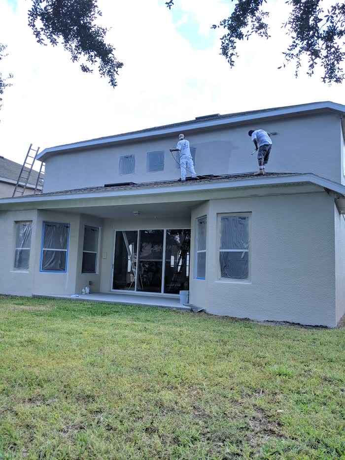 Two of the crew stand on a lower roof spraying the upper wall pale grey, windows masked off.