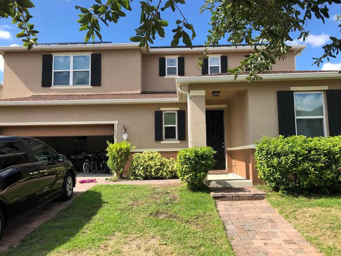 A tan two-storey house with dark shutters and a dark front door, hedges and a paver walkway out front.