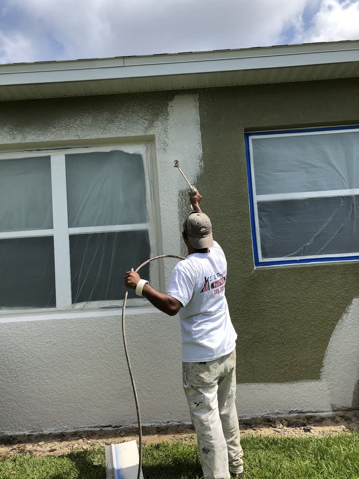 A worker aims a spray gun at a wall, olive green going on over pale grey beside a taped-off window.
