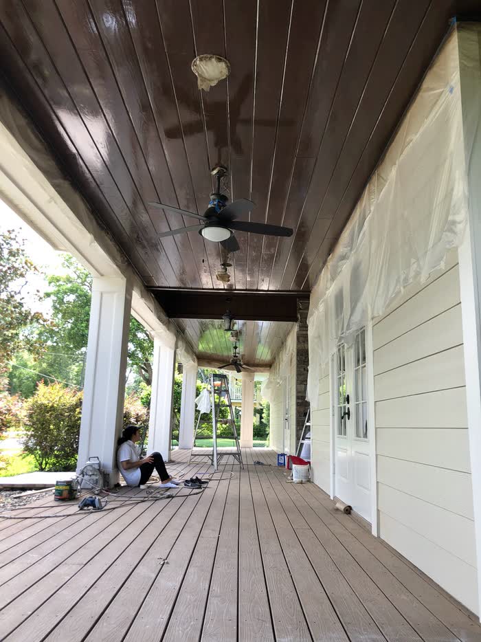 Long covered porch with a dark stained board ceiling, plastic sheeting over one wall, a worker resting on the decking.