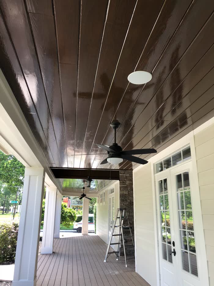 Dark brown porch ceiling boards, freshly coated and still glossy, above white columns and a black ceiling fan.