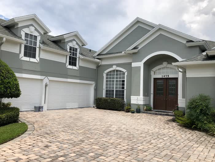 Grey-green two-storey home with white trim, twin white garage doors and an arched entry above a block-paved driveway.