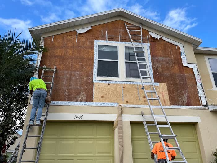 Two of the crew work on a house gable stripped back to bare board, ladders up against the wall.