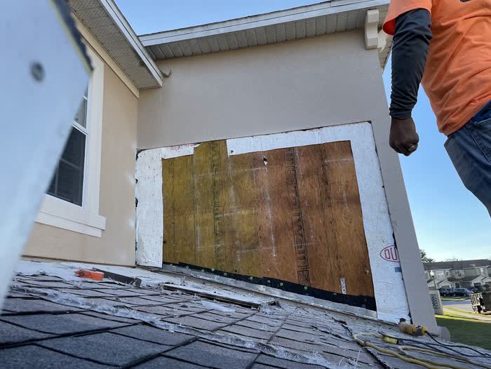 From a shingled roof, bare board fills a large opening cut into a tan wall beside a worker.