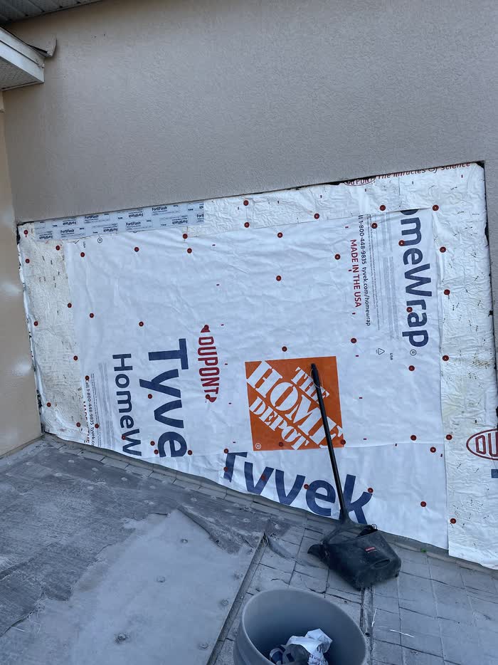 Taped white sheeting covers a low section of tan wall, a broom and bucket left on the paving.