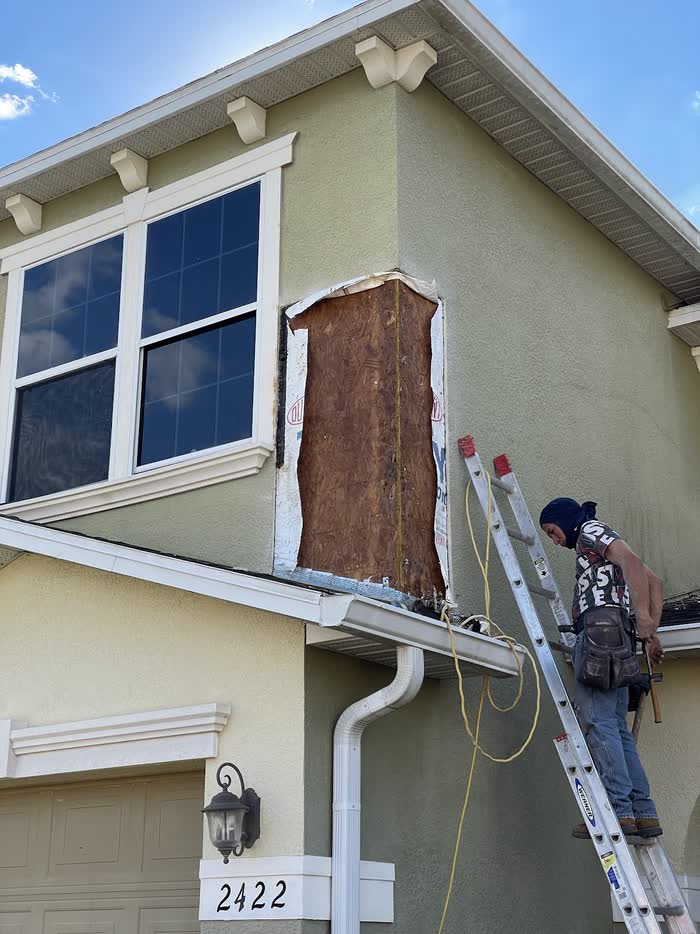 On a green house, a worker on a ladder reaches a tall narrow opening cut beside an upstairs window.