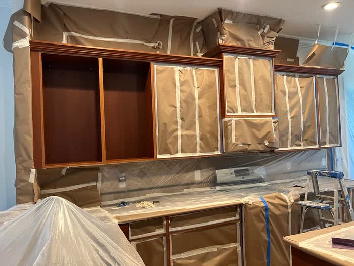 Reddish-brown wall cabinets with the doors taken off, openings papered and taped, counters sheeted in plastic.