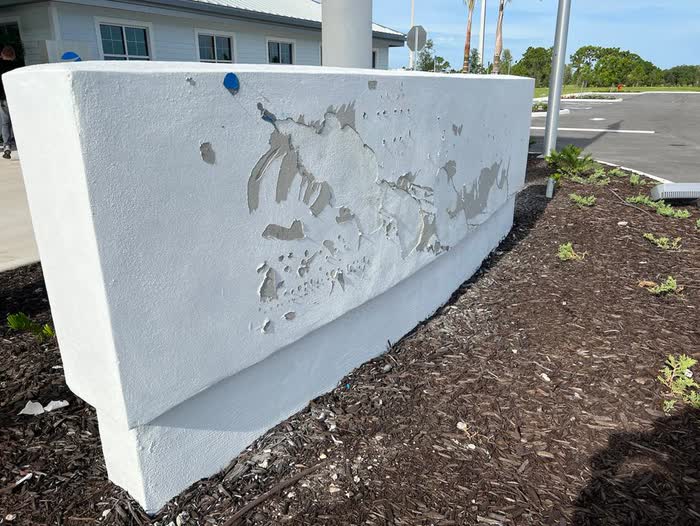 Flaking white coating on a low entrance wall, grey showing through, mulch bed and parking area behind.