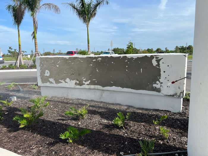A low white wall with grey patching spread across most of its face, palm trees behind.