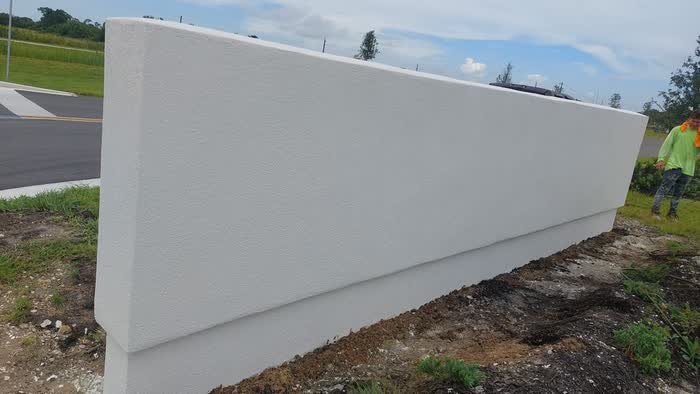 Long white wall running beside a road, a worker in an orange vest at the far end.