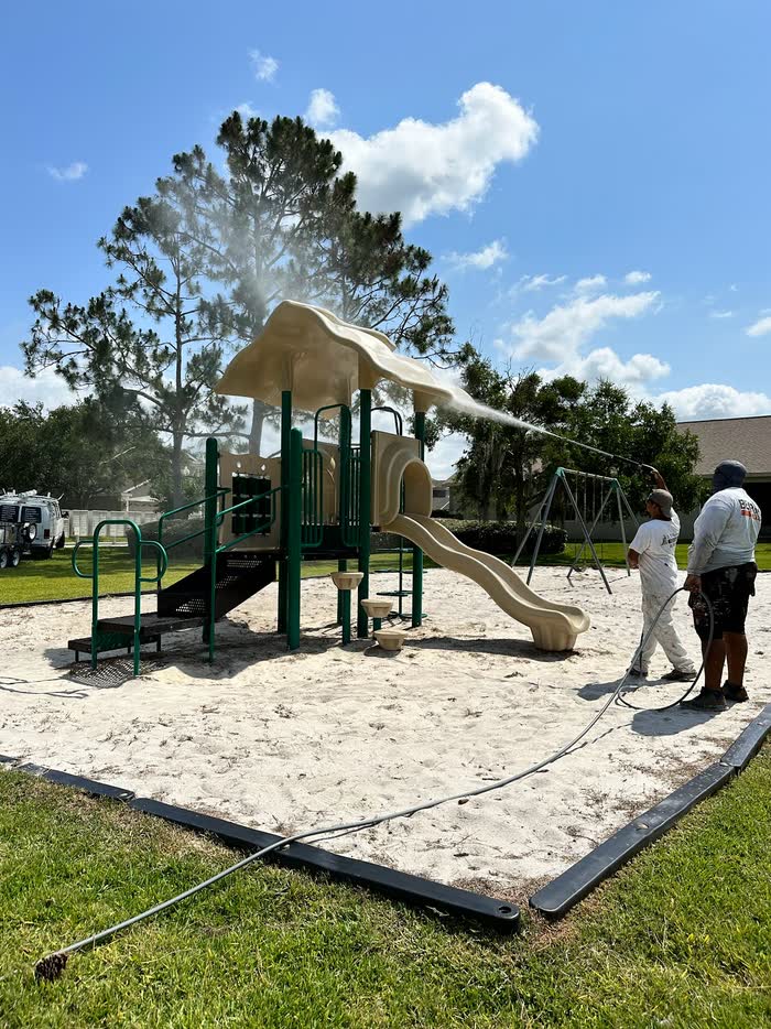 Two of the crew rinse a green and tan playground set, spray arcing over the slide onto sand.