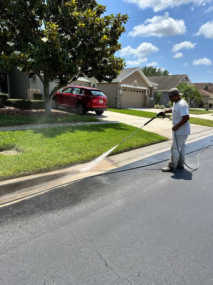 Standing in the road, a worker fans a spray along the curb, a wet line spreading ahead of him.