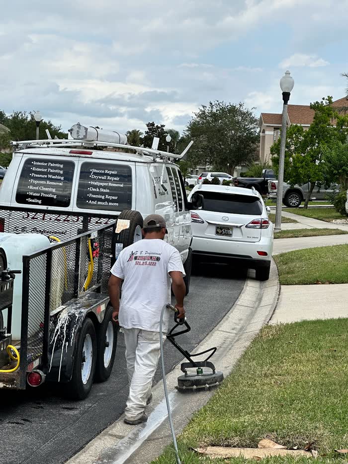 Beside a loaded work trailer and van, a worker guides a round scrubbing head along a wet sidewalk.