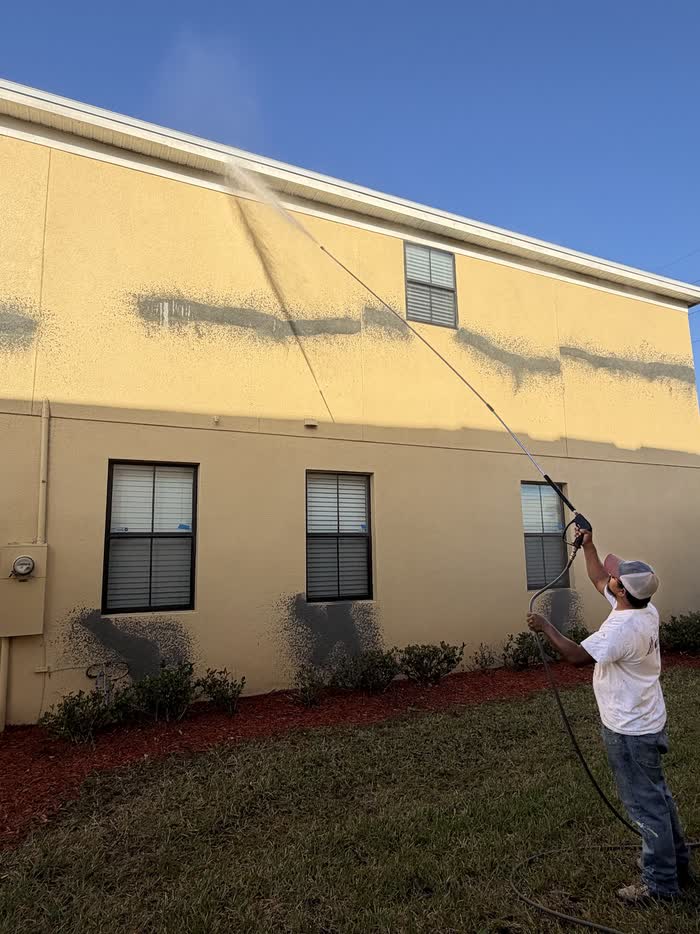 A worker sprays water from a long wand across a pale yellow two-storey wall streaked with grey grime.
