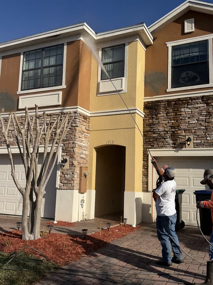 Water from a long wand arcs up toward the upper windows of a tan townhouse with a stone-look lower wall.