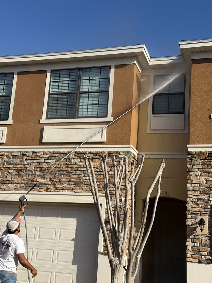 Spray arcs up to the eaves above a stone-look garage front as a worker steers the wand from below.