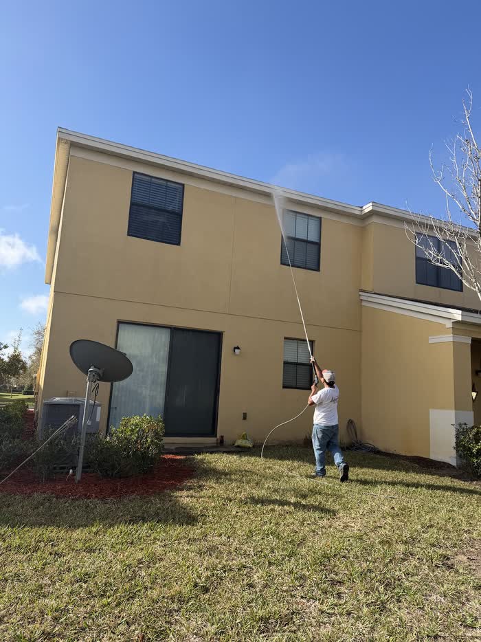 From the back lawn, a worker aims a long wand at the roofline of a yellow two-storey house.