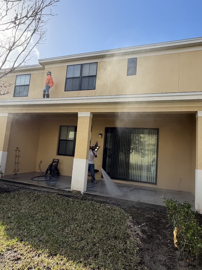 One of the crew washes a covered porch floor while another stands on the roof above, mist drifting across.