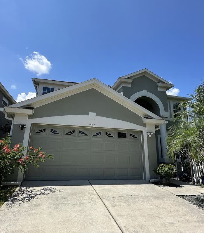 Front of a two-storey house in soft sage green, its garage door painted to match, under bright sun.