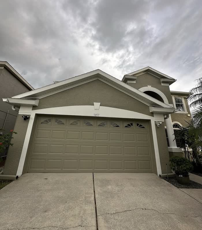 Olive-tan front elevation with a matching garage door and white trim, photographed under a heavy grey sky.