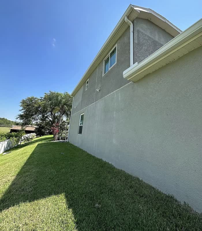 Side wall of a house in pale green with white gutters, seen along a sunlit stretch of lawn.