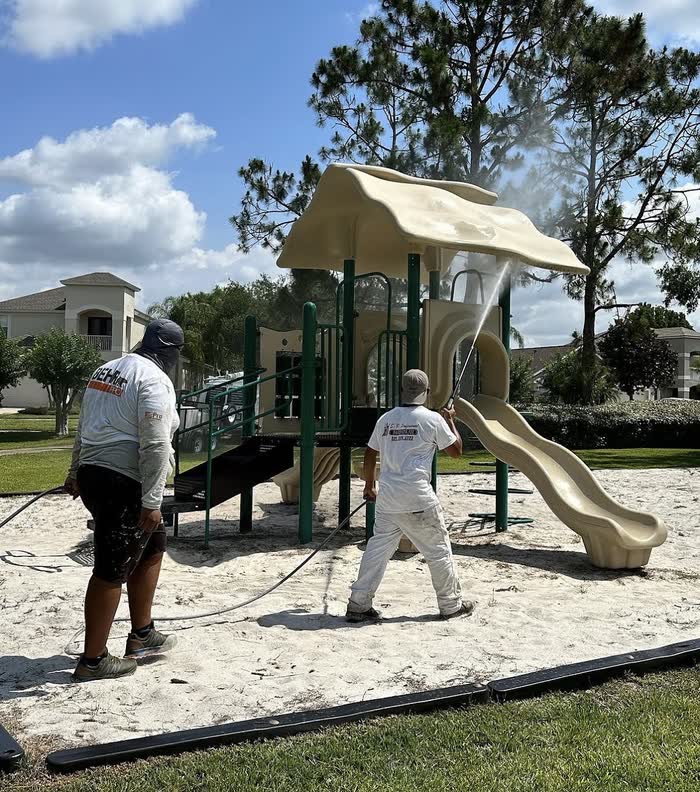 Two of the crew rinse a playground set, spray hitting the canopy above the tan slide and green posts.