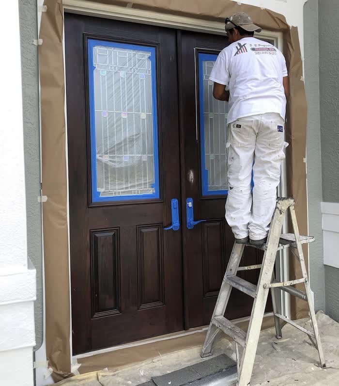A painter on a stepladder works on dark brown double front doors, glass masked with blue tape.