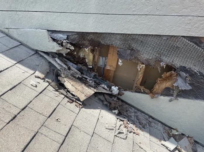A hole torn in a grey wall just above a shingle roof, with debris scattered below.
