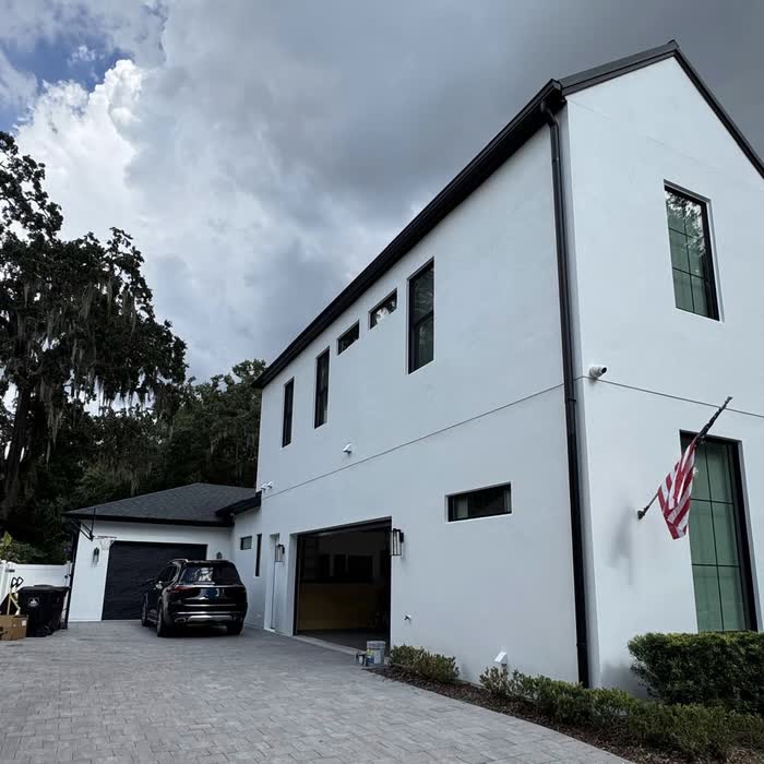 White two-storey house with black trim and gutters, a car in the paver driveway under storm clouds.