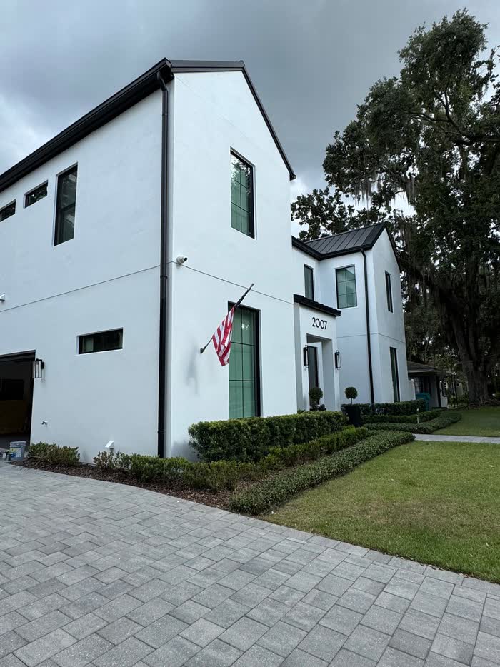 Two-story white home seen from the driveway, black trim and gutters, with a flag above a clipped hedge.