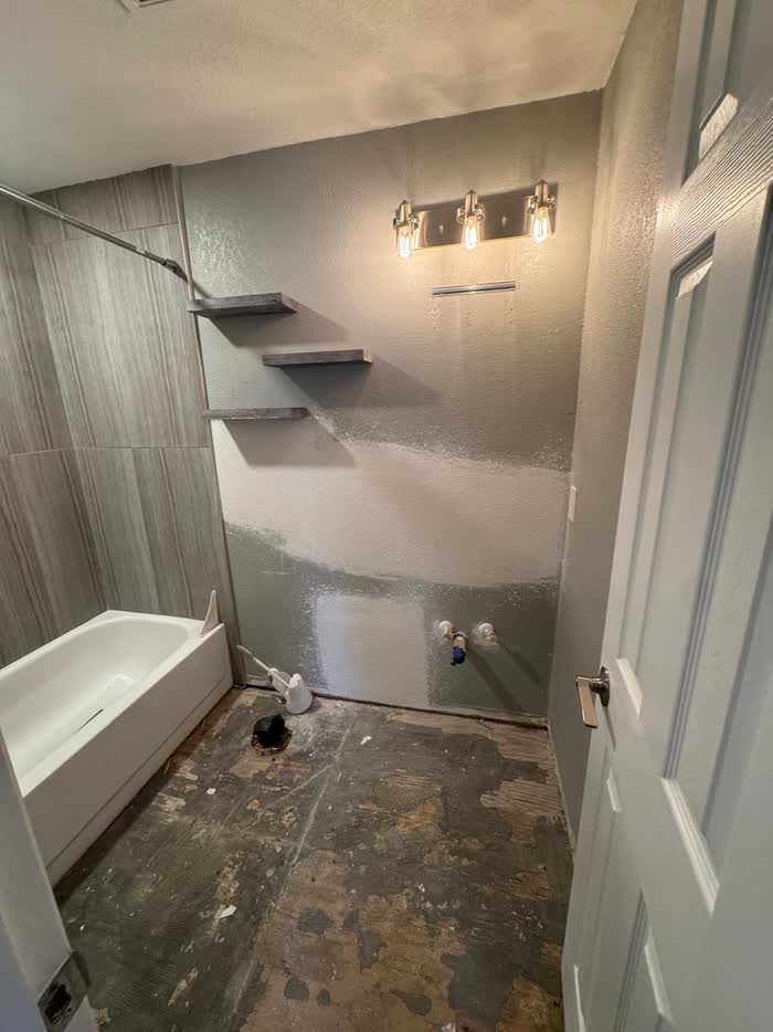 Grey bathroom walls mid-repair behind a white tub, with three floating shelves and a bare stripped floor.