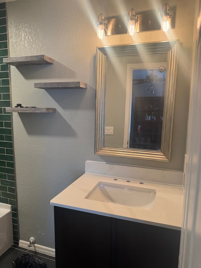Dark vanity with a white top, framed mirror and three-bulb light beside three stacked floating shelves.