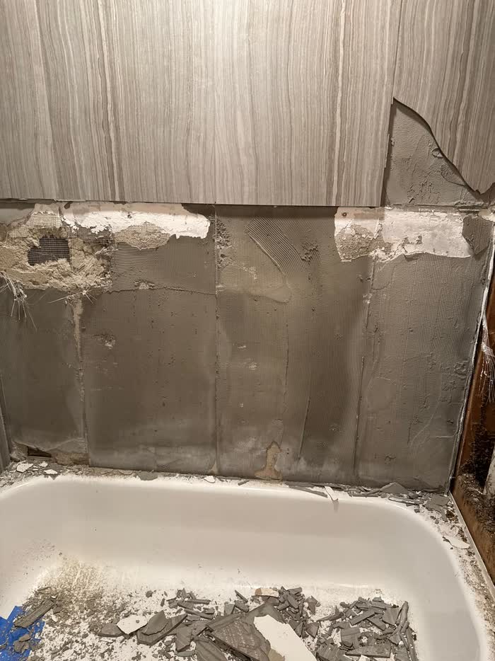 Wall tiles stripped back to grey backing above a tub, with broken pieces heaped inside.