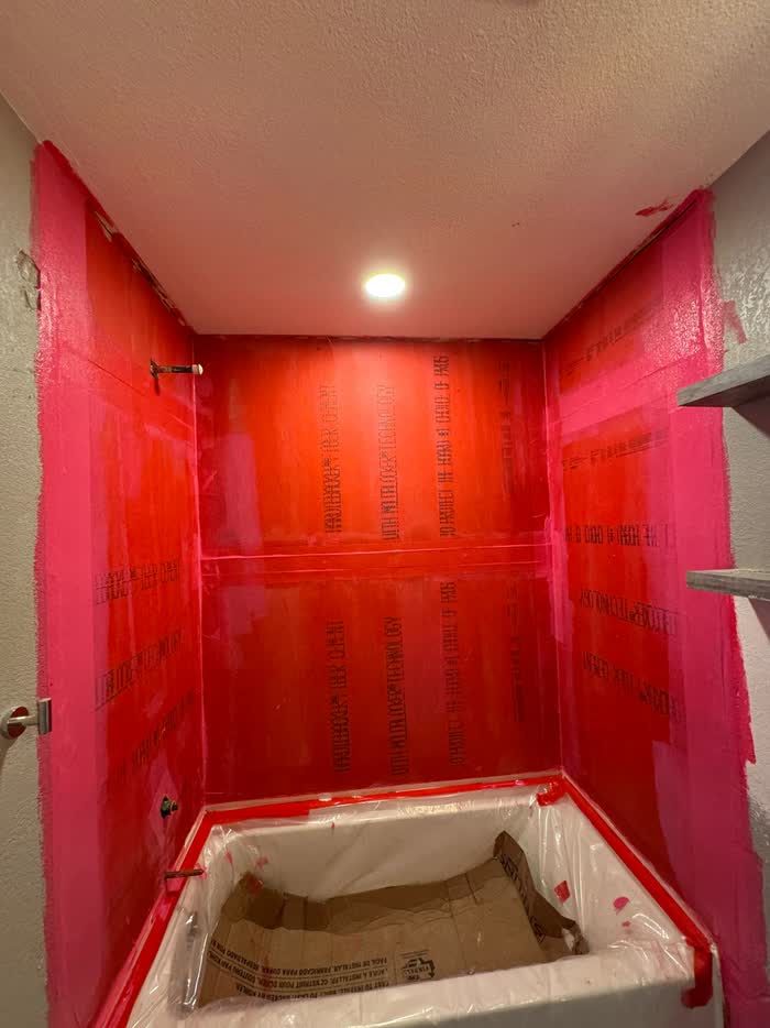 Red waterproofing coats the three walls of a narrow shower alcove, its base masked with plastic.