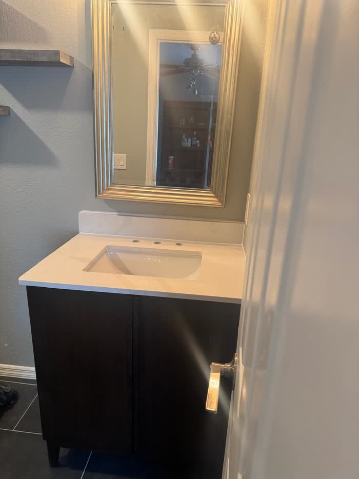 Dark vanity cabinet with a cream countertop and undermount basin below a framed mirror on a grey wall.