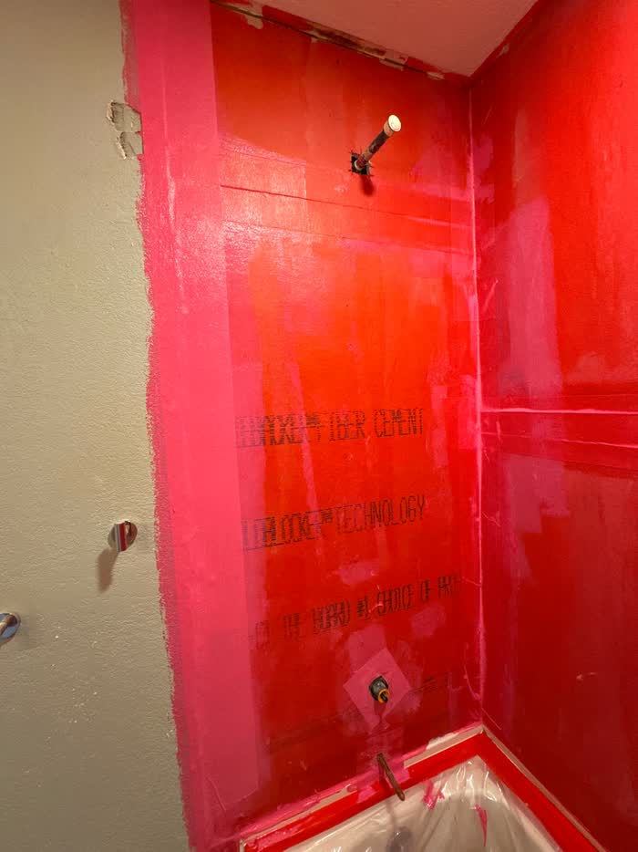 Bare bulb and a capped pipe stub stand out against red-coated shower walls, plastic sheeting below.