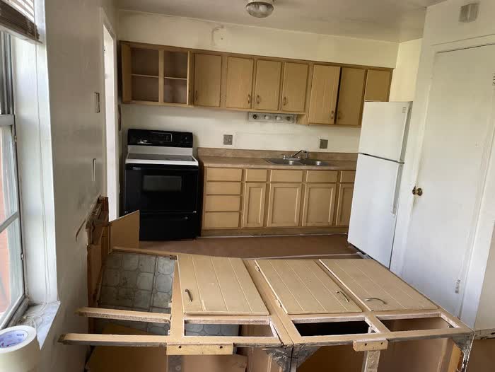 Older kitchen with pale tan cabinets, a black range, and a peninsula stripped back to its frame.