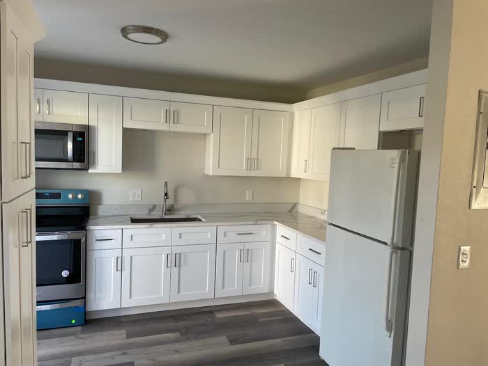Corner view of a white kitchen with a fridge, new range and grey plank flooring.