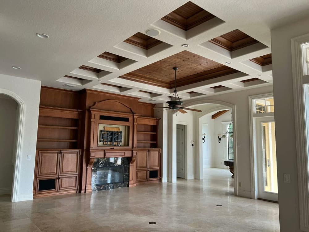 A living room with a coffered ceiling in white and stained panels, built-in shelving either side of the fireplace, and arched openings through to the rest of the house.